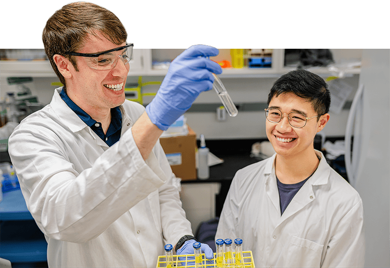 Two researchers in lab equipment examine a vial with solution.