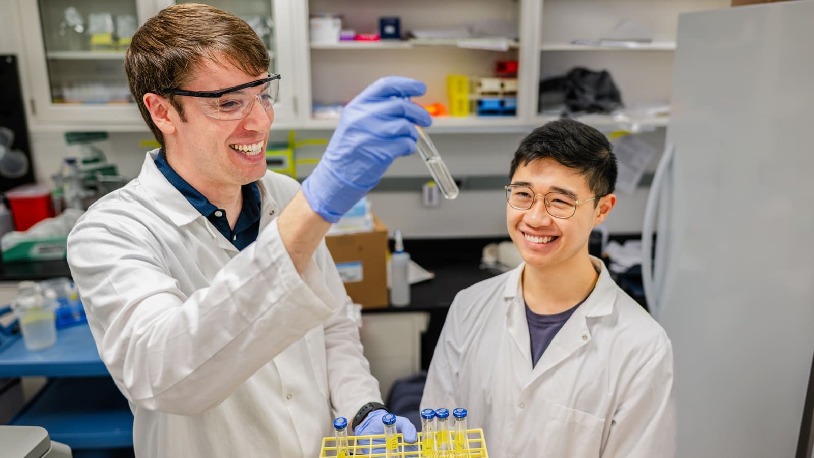 Two researchers in lab equipment examine a vial with solution.