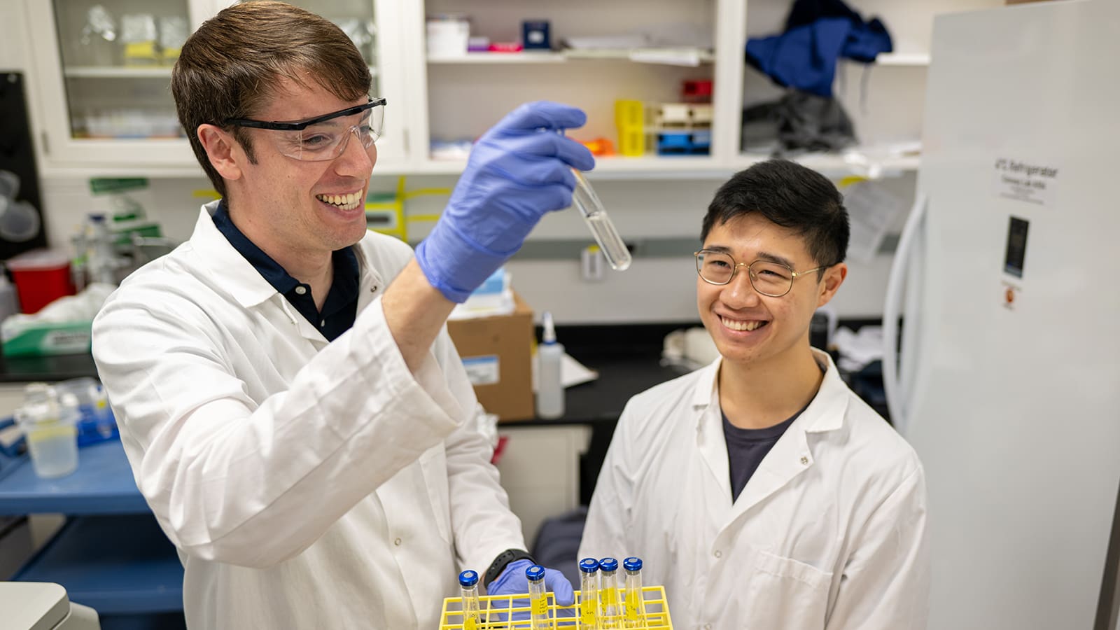 Two researchers in lab equipment examine a vial with solution.