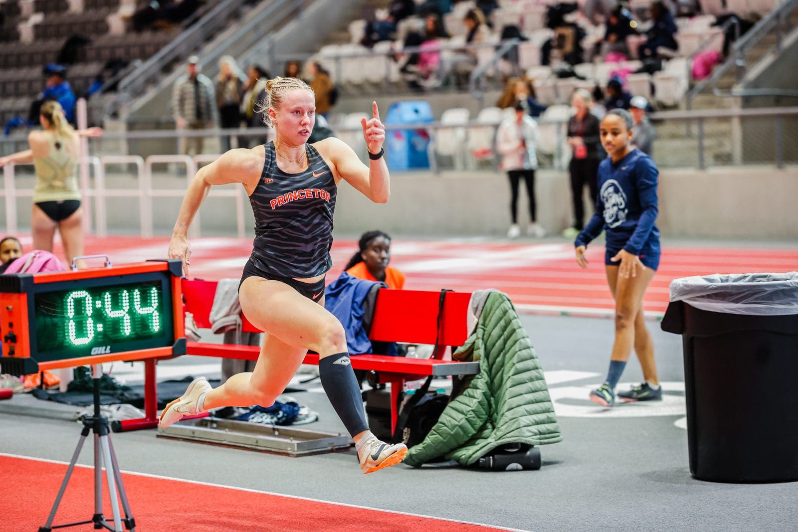 A woman runs on a track. A large digital timekeeping sign reading "0:44" is visible on her right side.