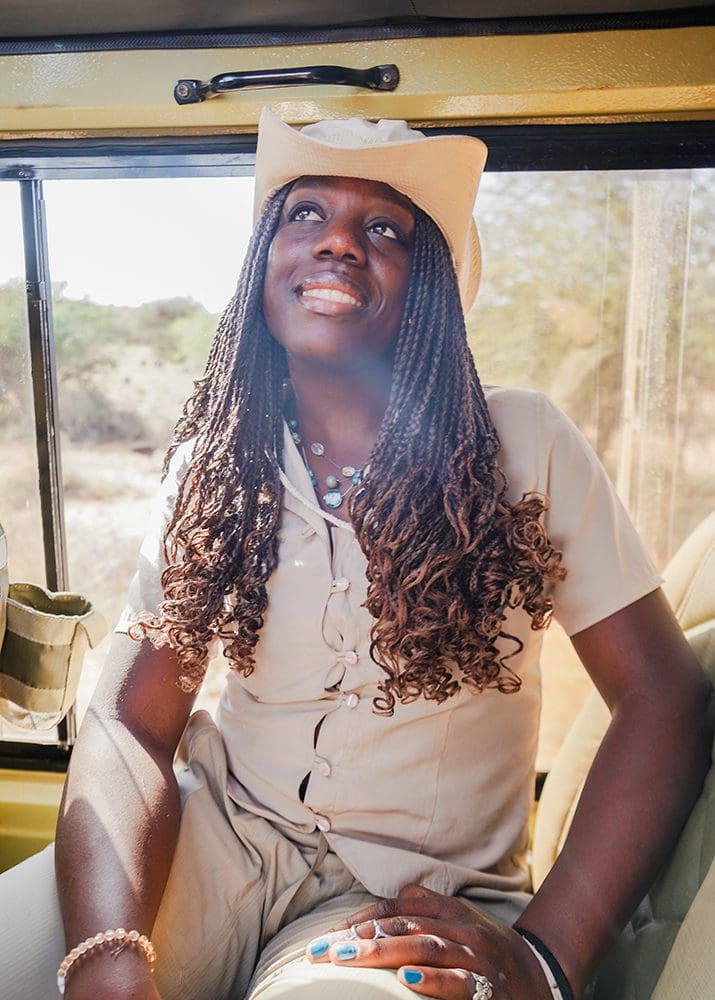 A woman wearing a tan hat and shirt sitting in a vehicle, her back to the window. She is gazing up.