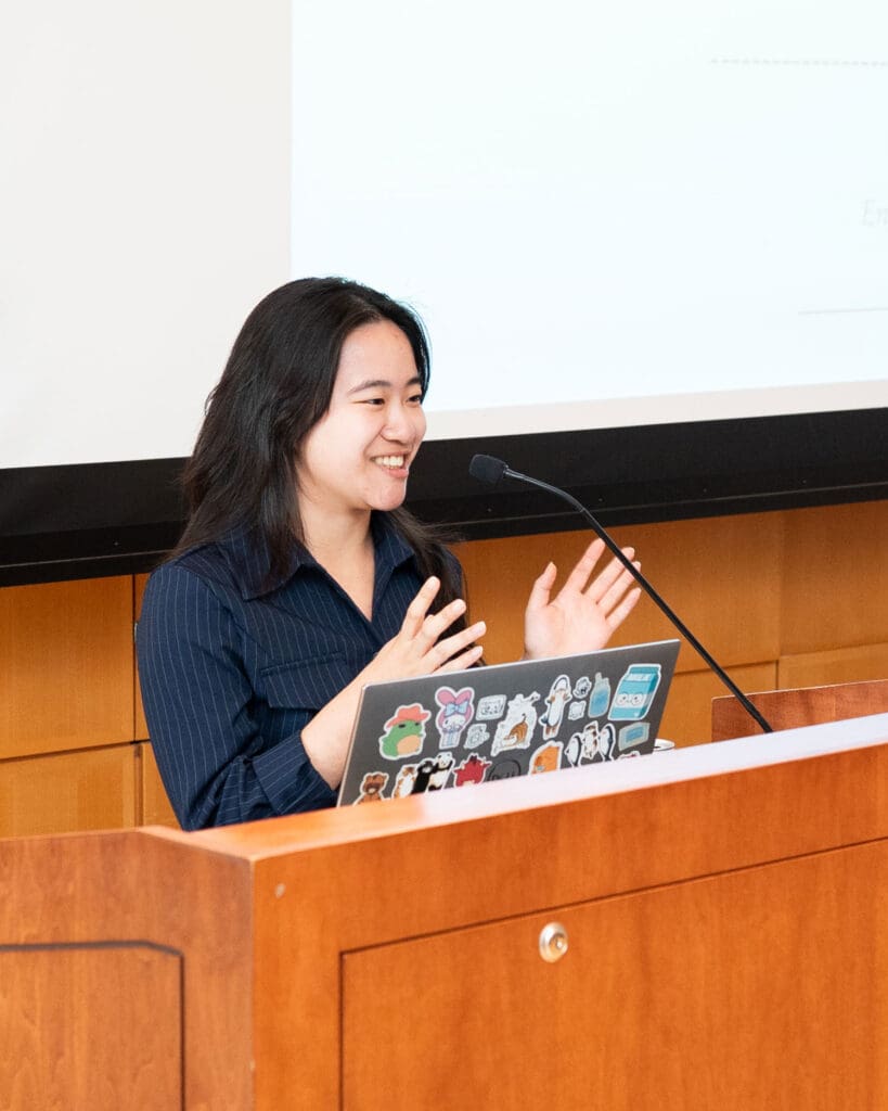 A woman standing at a podium with a laptop, speaking to an audience.