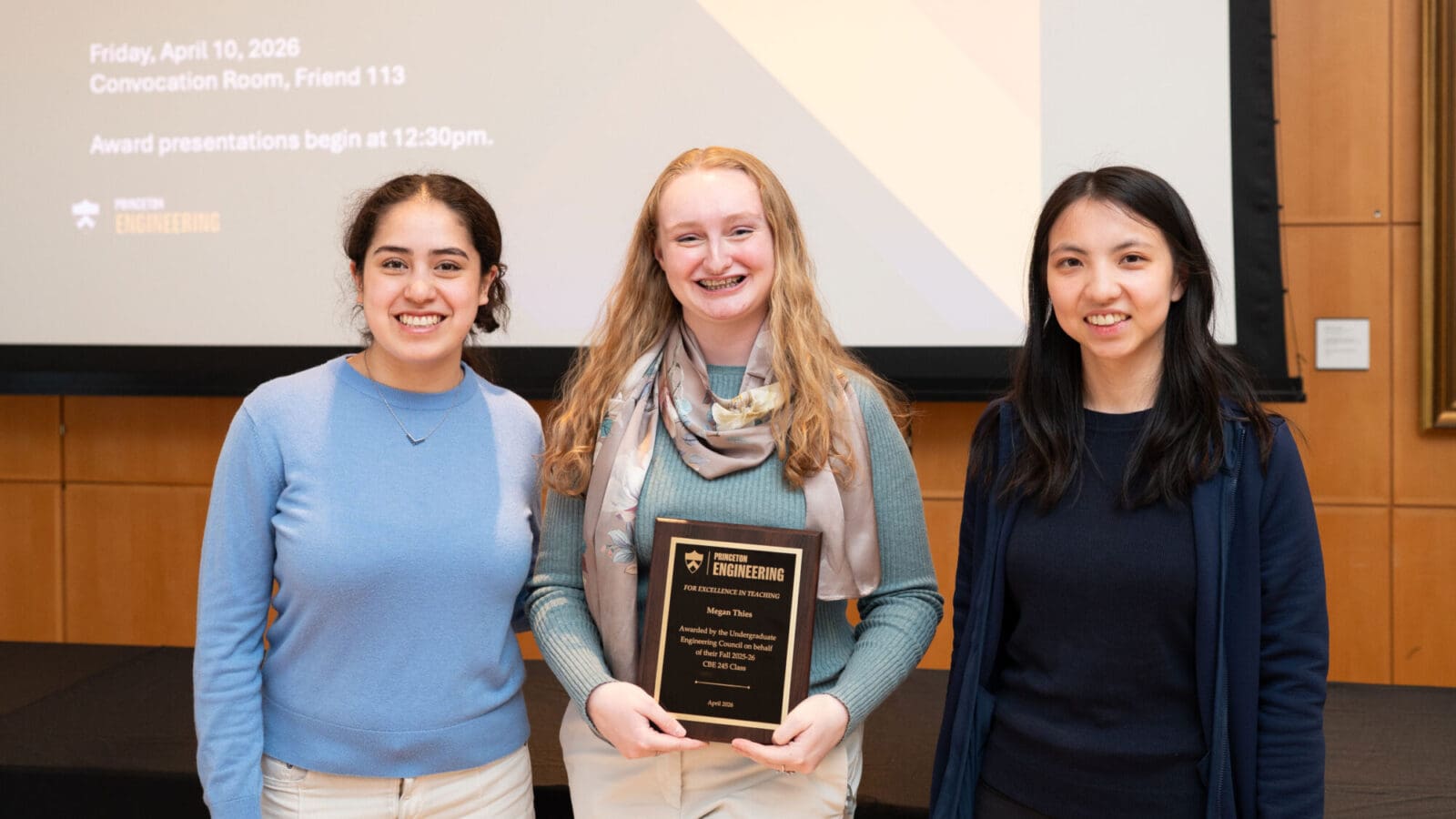 Three women stand together; the woman in the center is holding an award plaque.