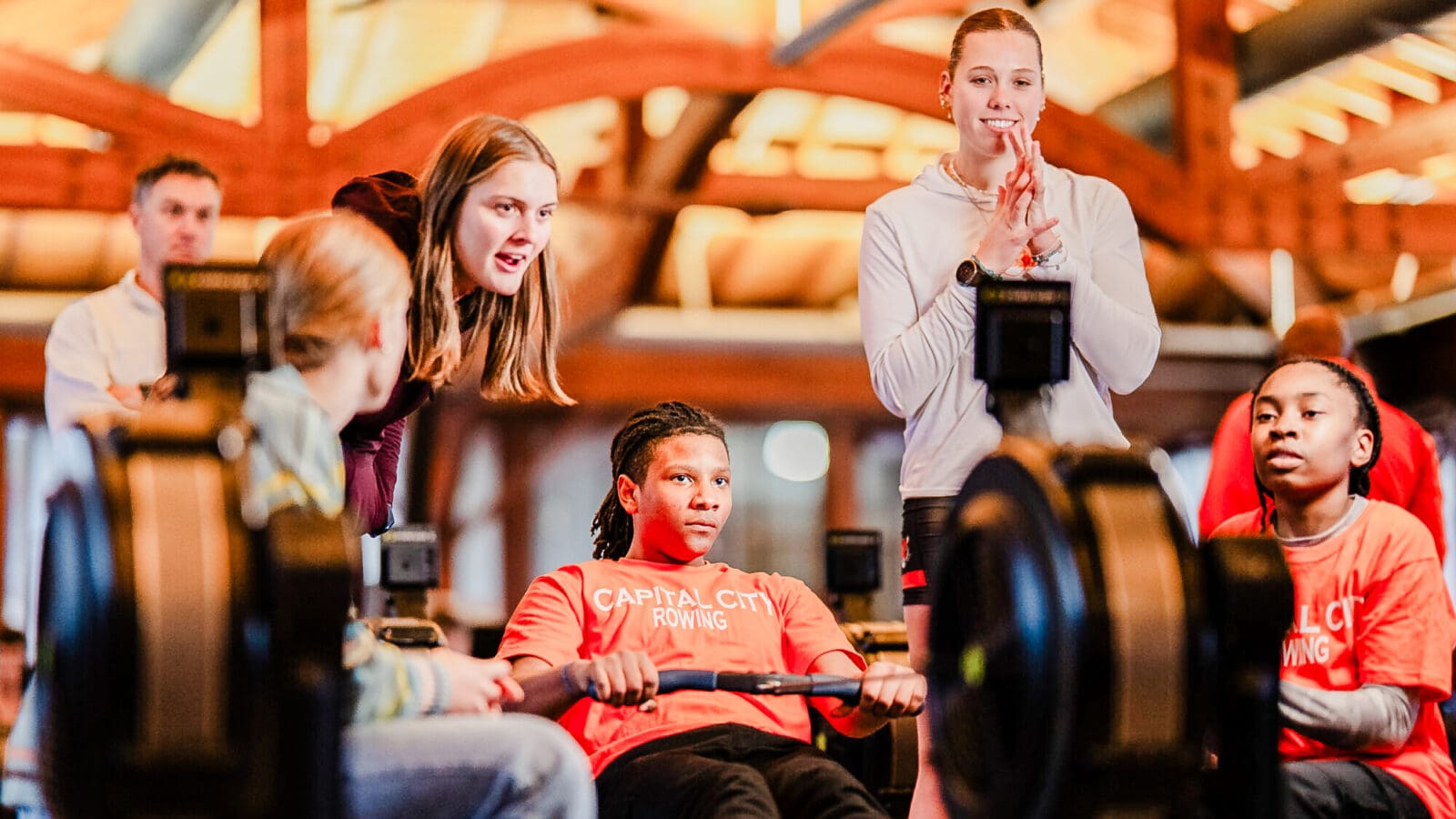 A middle schooler uses a rowing machine while two coaches, standing, cheer him on. Another middle schooler sits nearby.