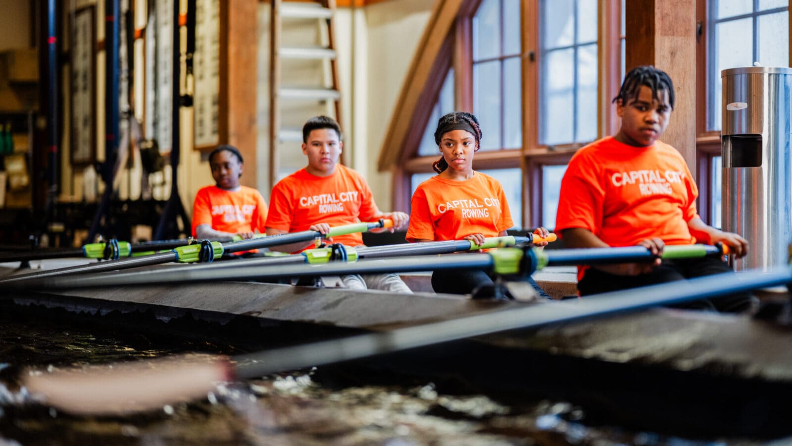 Four middle school students in orange shirts rowing together in an indoor crew training tank.