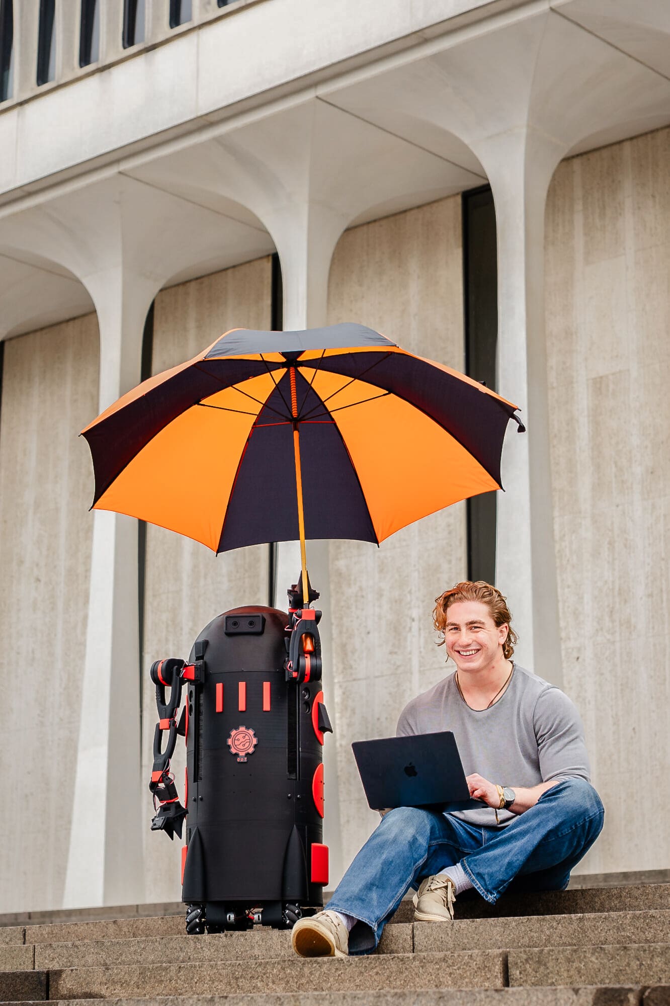 A man sitting on steps smiling with a laptop. A robot next to him holds an umbrella.
