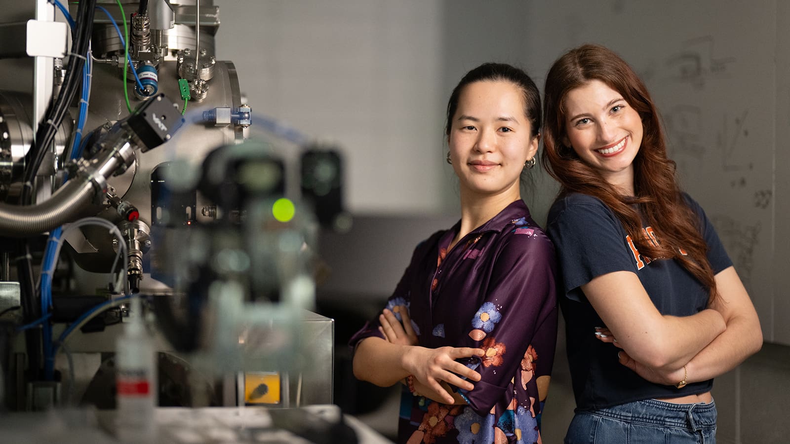 Laura Futamura and Kalli Zervas, quantum Ph.D. students, lean against each other in a high-tech lab.
