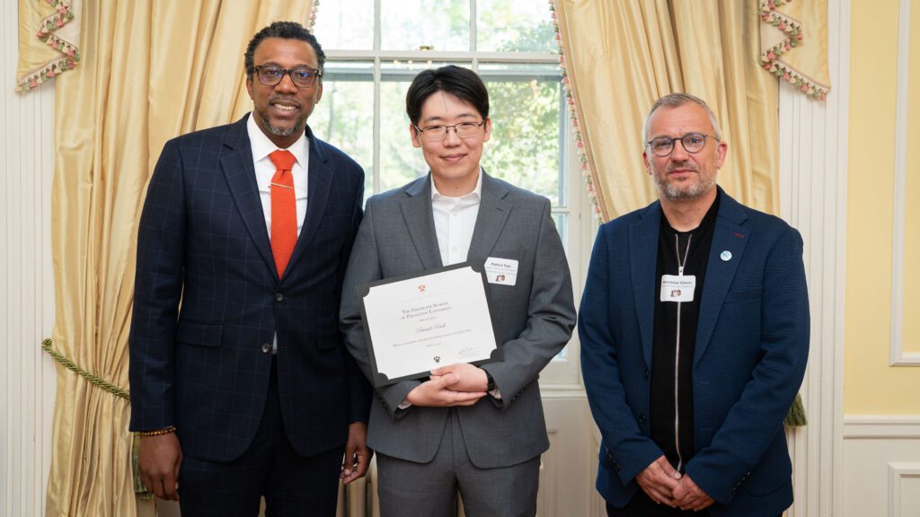 Three men in suits stand together, smiling at the camera. The man in the center is holding a certificate.