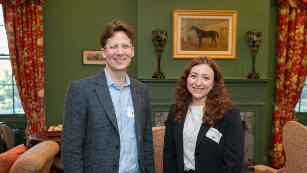 Two smiling people pose in a formal living room. Above the fireplace behind them is a painting of a horse.