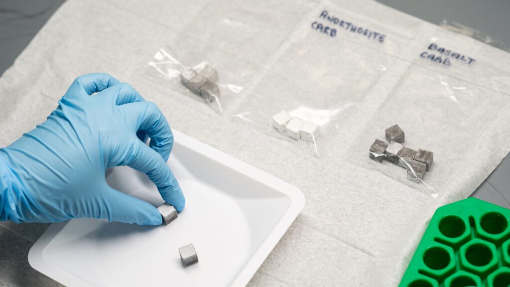 A person's hand in a blue laboratory glove places a small metal cube into a weigh boat. Also visible on the table are three clear plastic bags containing several metal cubes each. The cubes in each bag are a different shade of gray.
