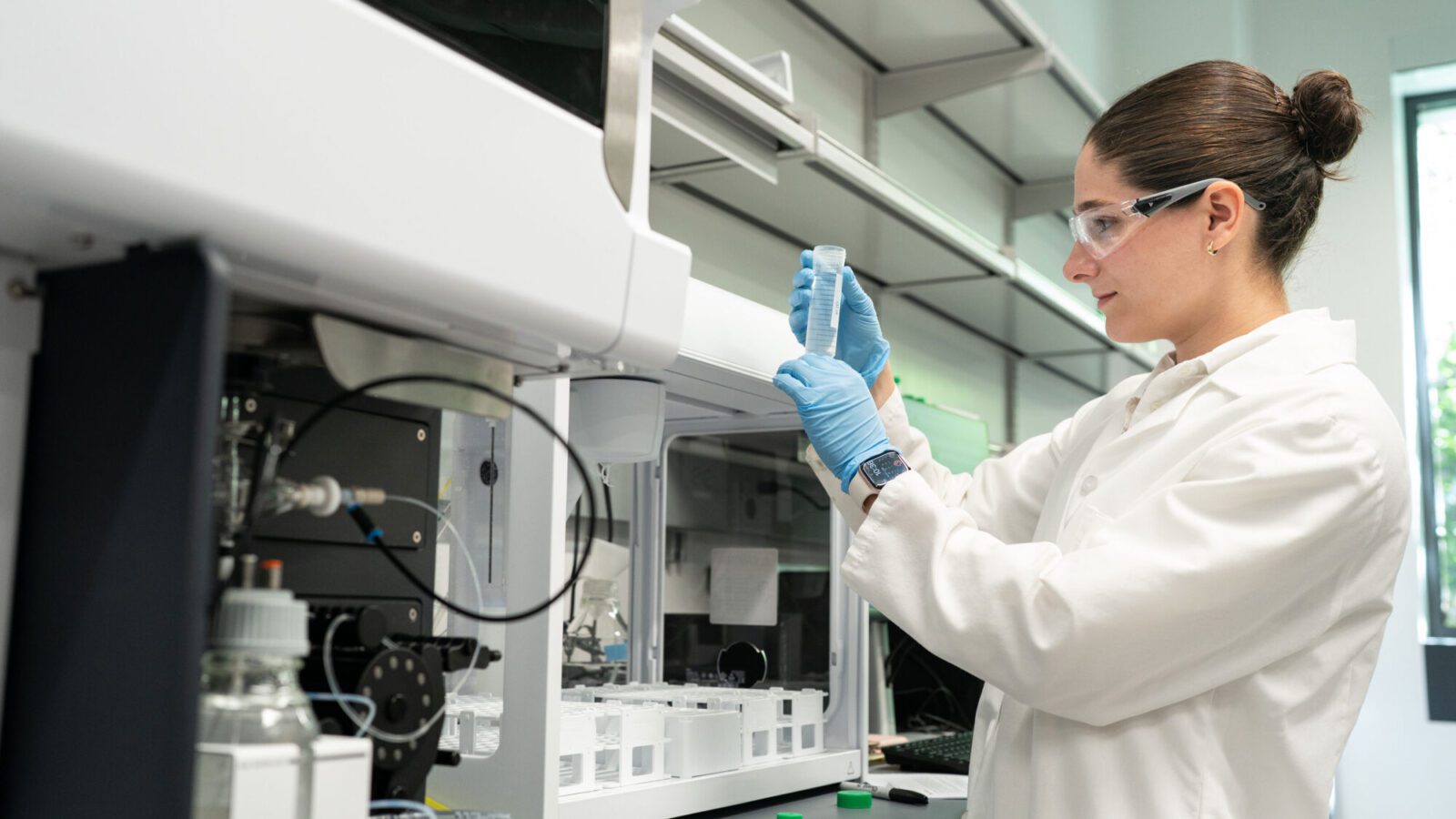 A woman in a lab coat and safety goggles prepares to place a test tube into a machine for analysis.