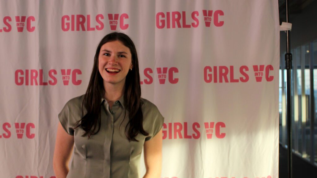 Student stands in front of banner printed with repeated logo of Girls Into VC.