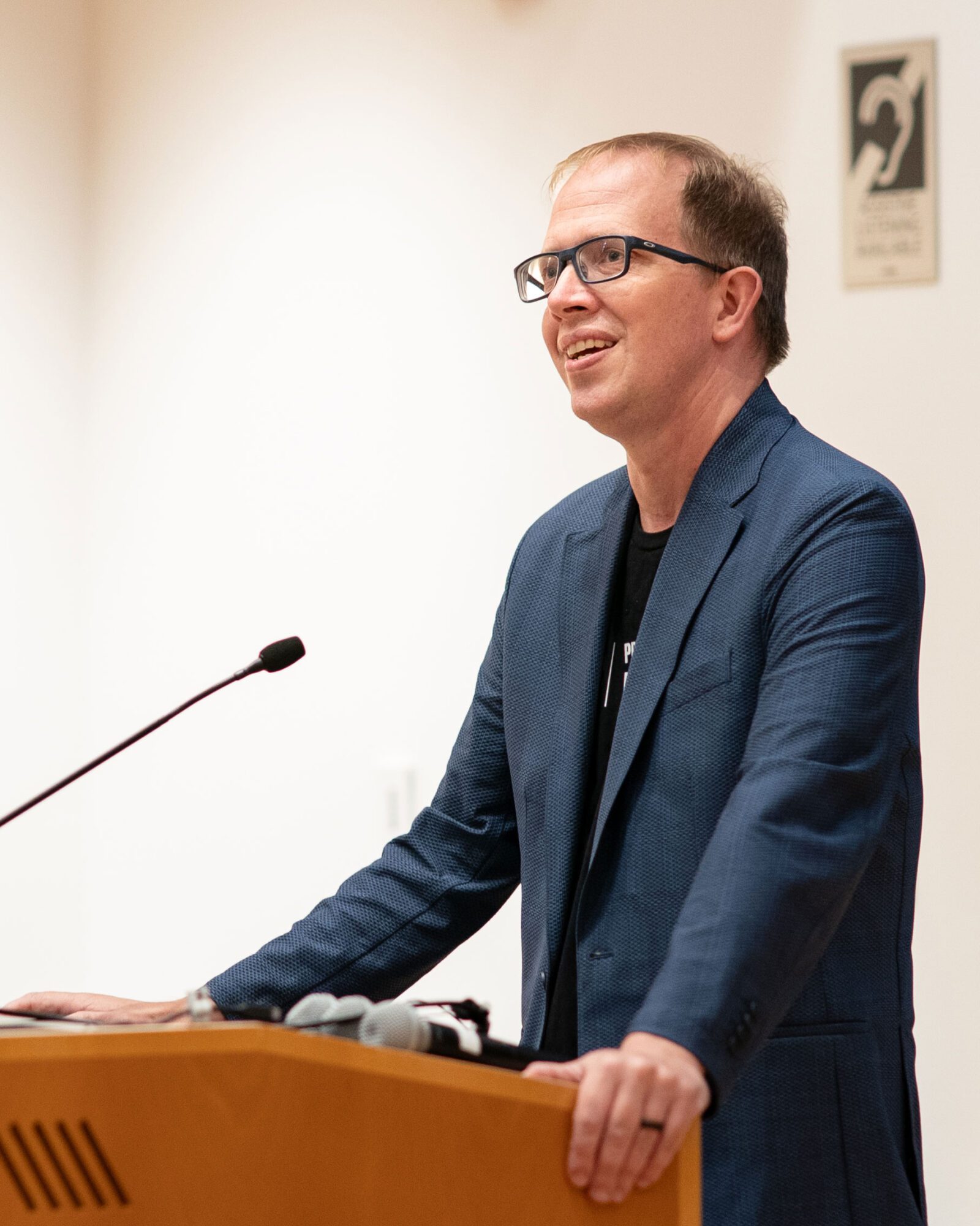 A man in a suit and glasses addresses an audience at a podium.