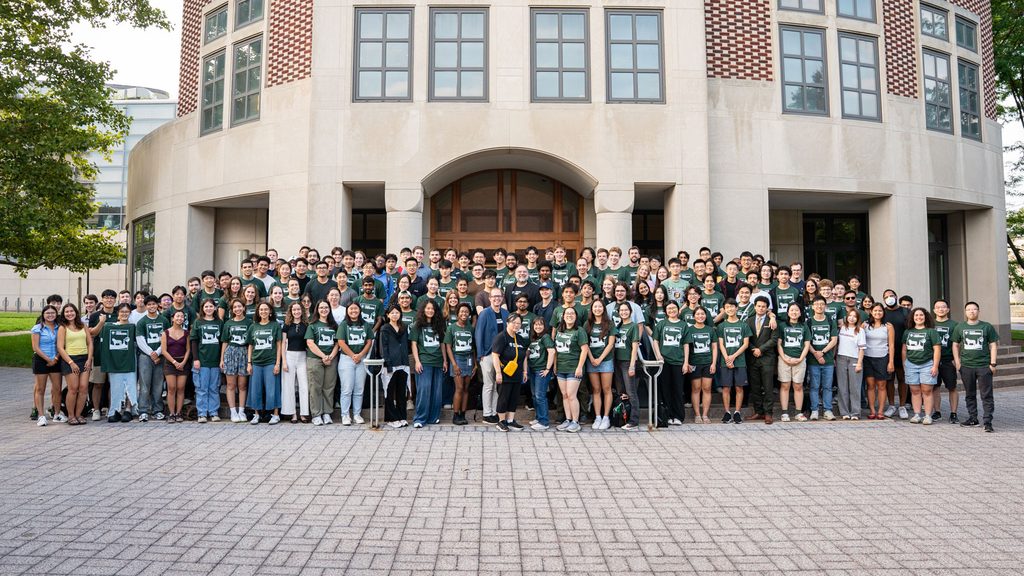 A large group of individuals, many wearing green shirts, stands together in front of a building, smiling and posing for a photo.