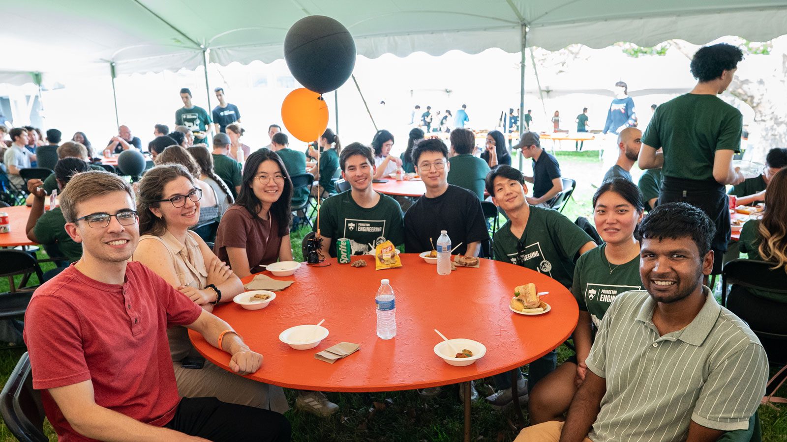 A group of eight students seated around a table under an event tent.