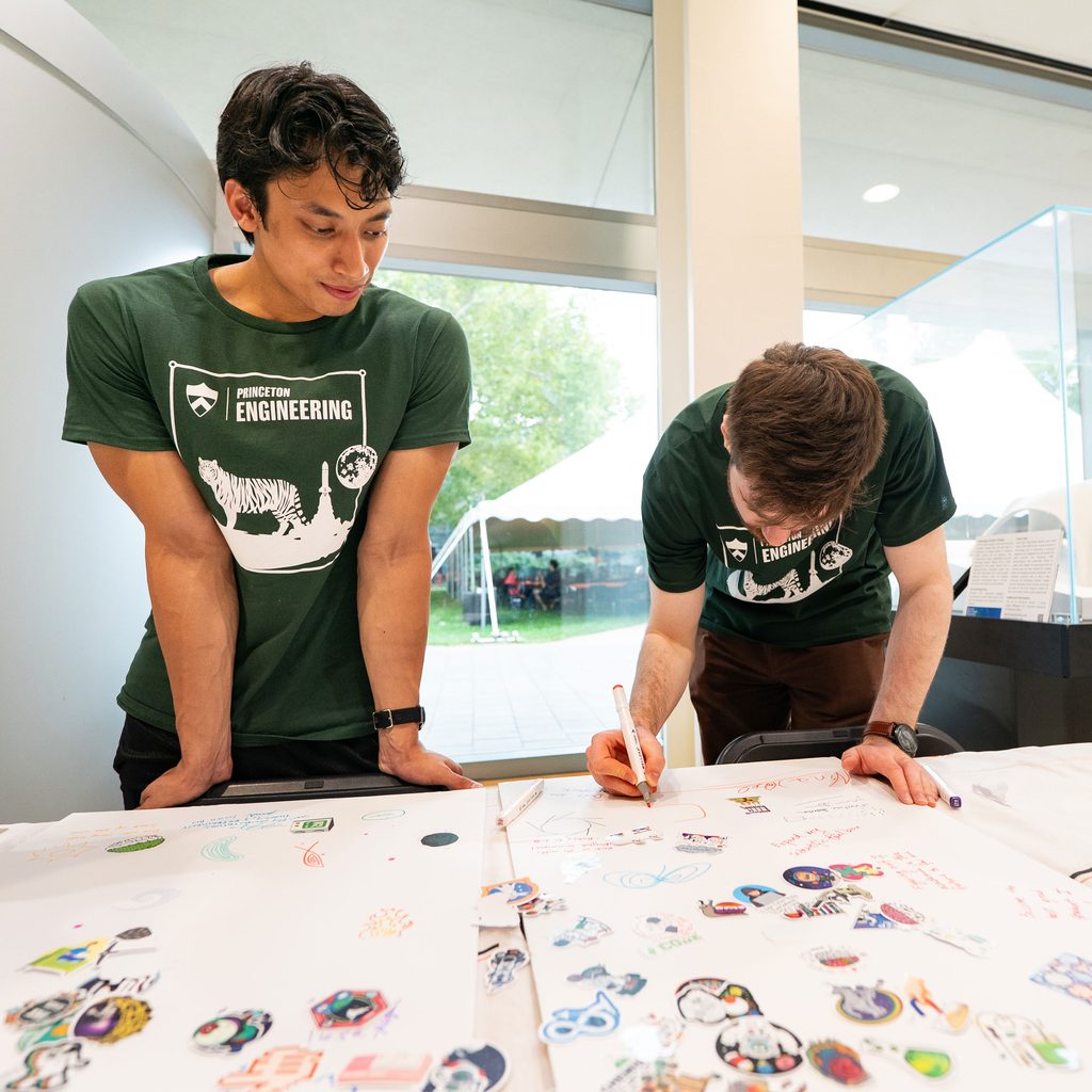 Two students in green Princeton Engineering T-shirts stand at a table with white posterboards and stickers on it; one of the students is leaning down and writing on one of the posterboards.