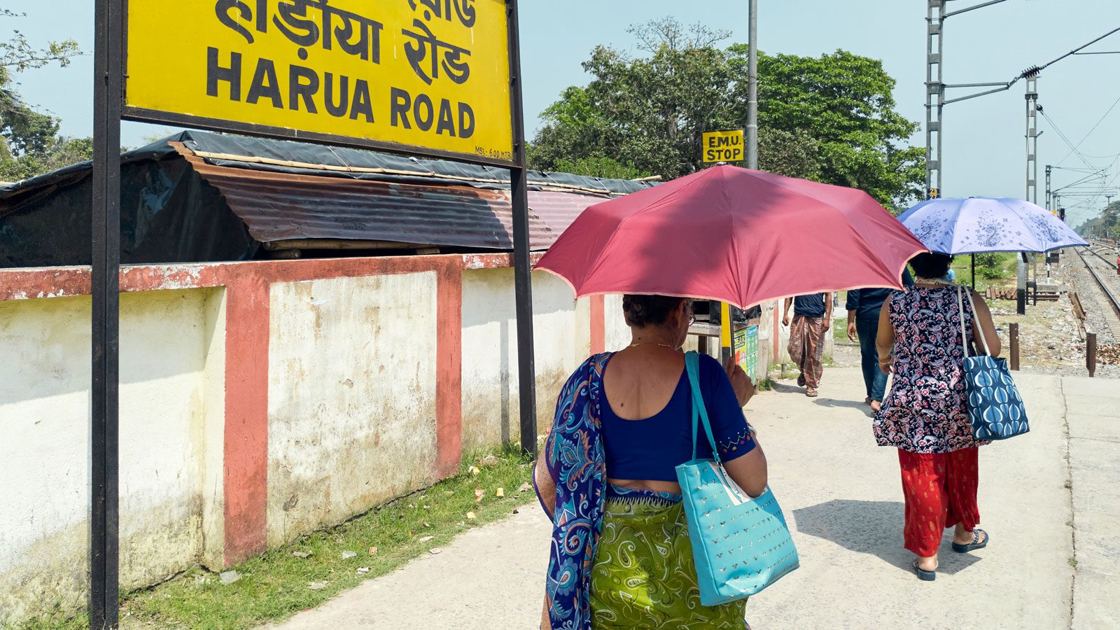 Two women holding umbrellas walk along a train platform on a sunny day. They are facing away from the camera.