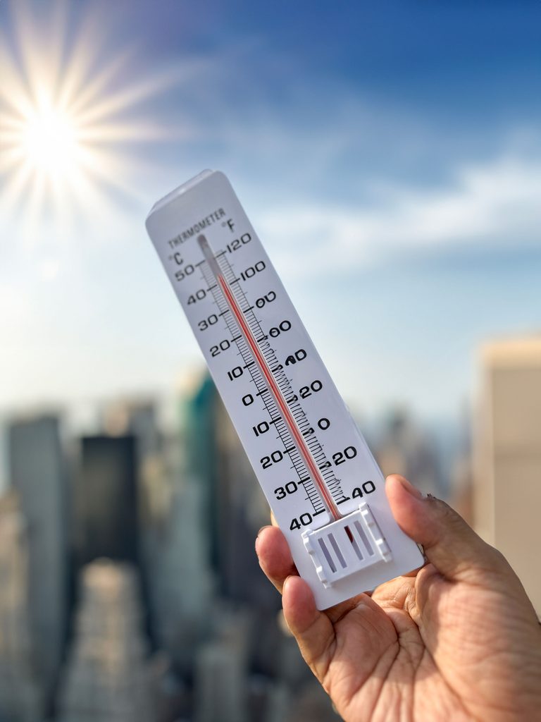 A hand holds a thermometer in front of a city skyline. The temperature reading is above 40 degrees Celsius (100 degrees Fahrenheit).