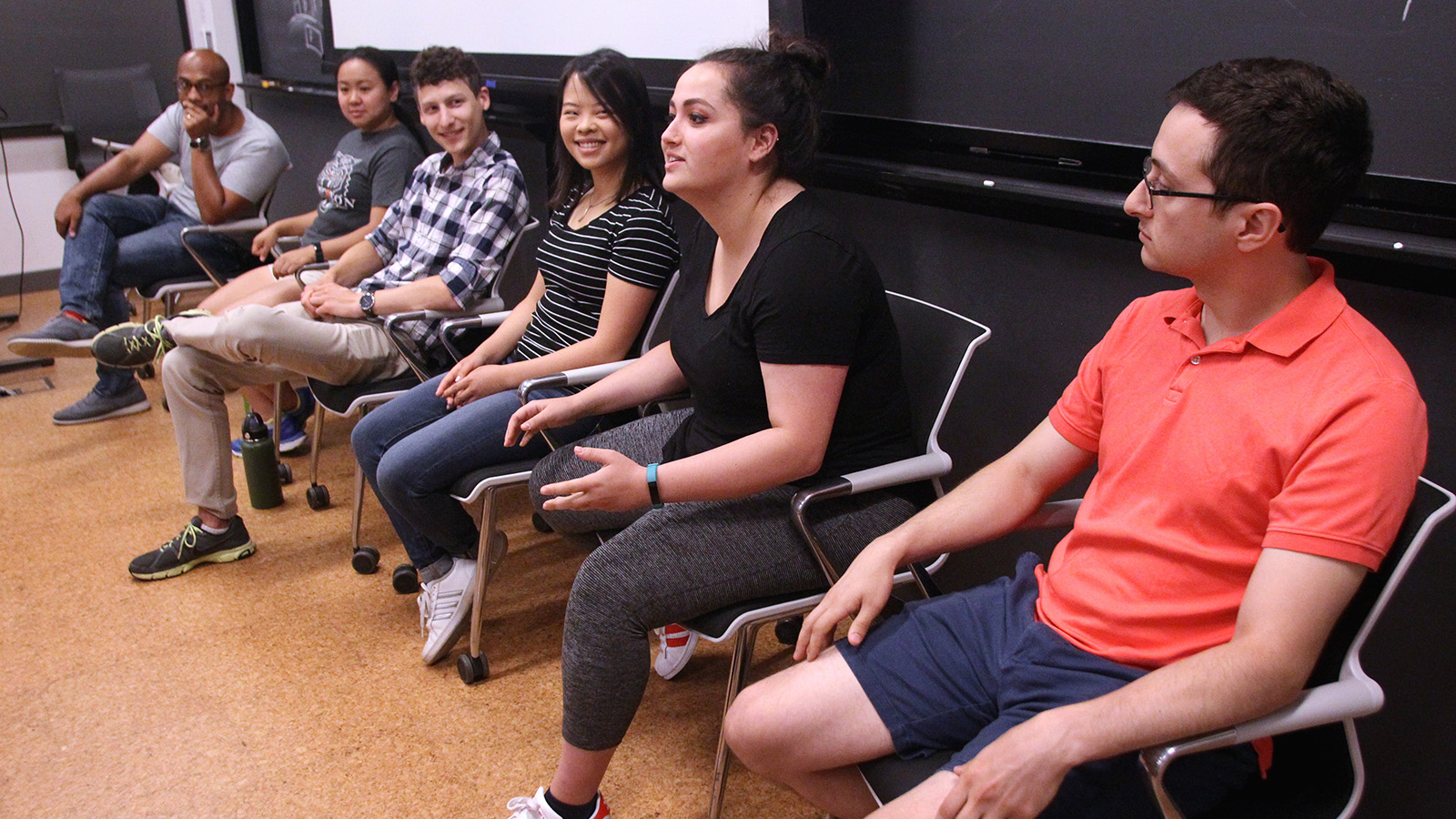 Five students and a professor sit in a row in chairs along a wall, shown from angle so the row of people recedes toward background from right to left.
