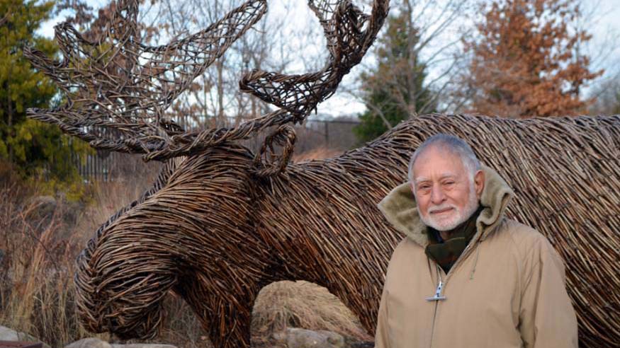 Robert Mark, standing in front of a Moose statue made of some kind of intertwined wood.