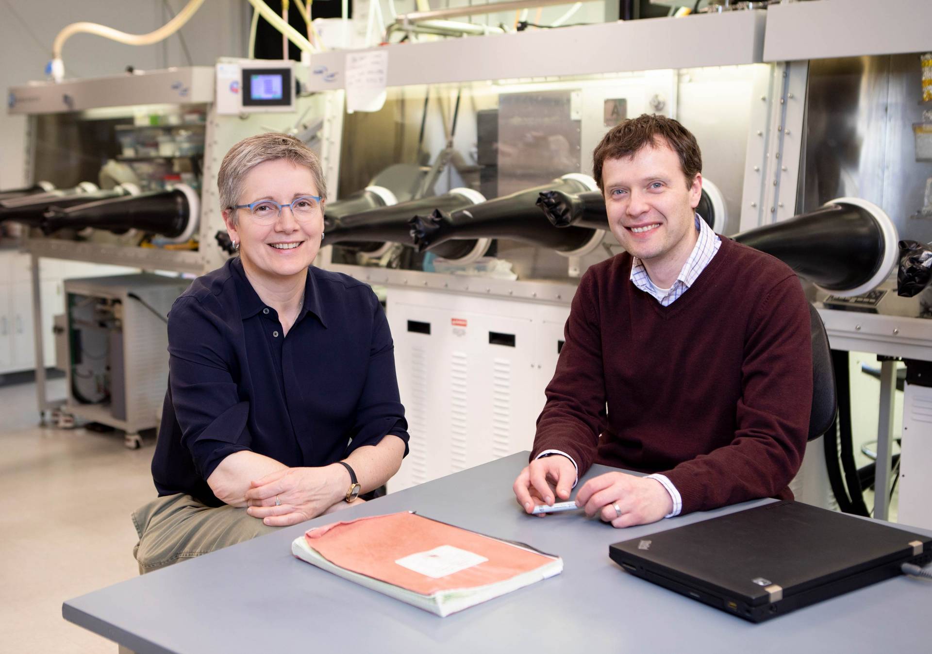 Two scientists, a woman and man, sit at table in lab and pose for photo